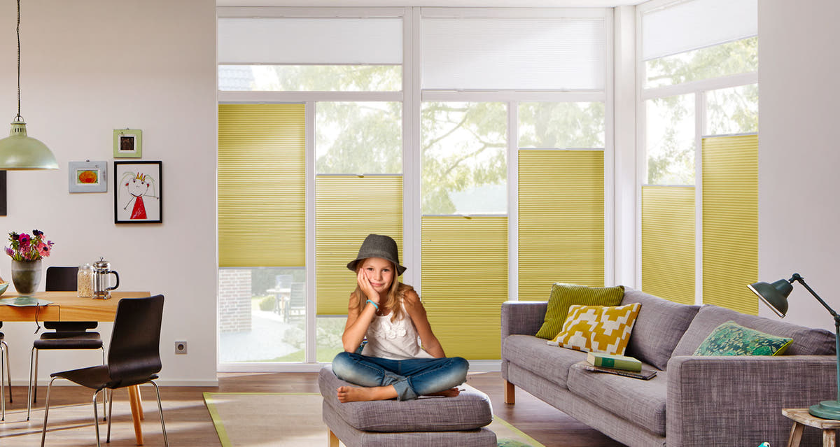 Wohnzimmer mit gelbgrünen Plissees, grauem Sofa und Sitzhocker